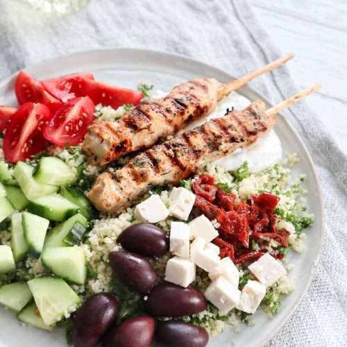 Assiette avec un fond de couscous garni de brochettes de poulet, de feta, de concombre, tomates, olives noires et tomates séchées