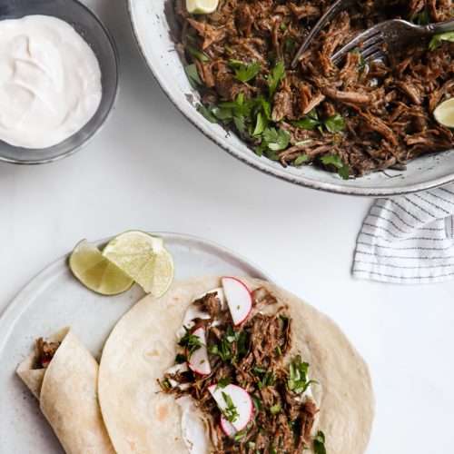 Un plat de service avec la barbacoa et quelques limes, une assiette avec deux tacos et un petit bol de crème sûre