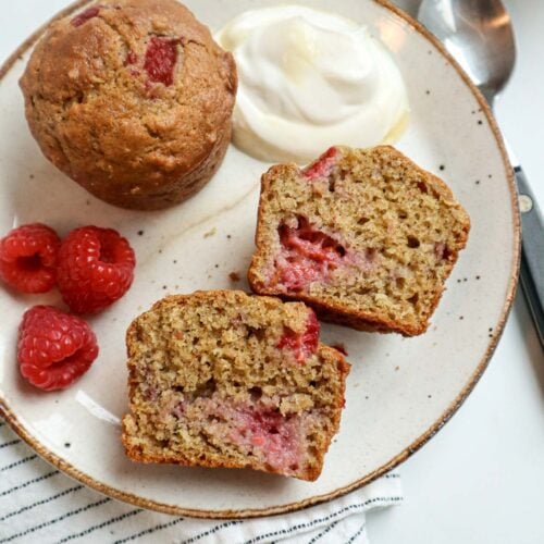 Muffins aux framboises coupé en deux déposé sur une assiette, accompagné de yogourt et de sirop d'érable et de quelques framboises fraiches