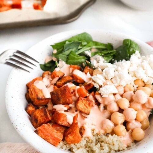 Un bol avec du quinoa garni de cubes de patate douce grillée, avec des pois chiches, des épinards hachés, un peu de grains de fromage feta. Sur le dessus, un filet de sauce d'apparence crémeuse.