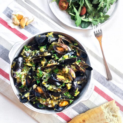 bol de service rempli de moules cuites napées d'une sauce poireaux et vin blanc, accompagné d'une salade verte et d'une baguette entamée